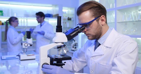 young student researcher man work with lab microscope for a research project while medical team talking in hospital laboratory room