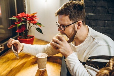 young man eat fast food and watch video on smartphone in cafe