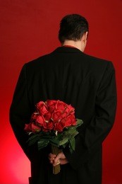 man holding out large bouquet of red roses behind his back on red backdrop