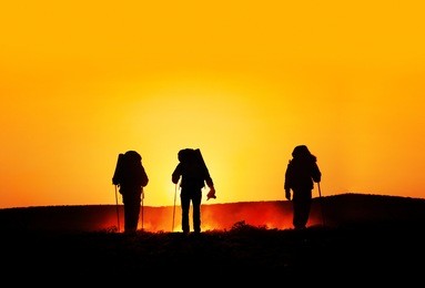 three tourist silhouettes walking on the hills with track sticks and backpacks at sunset orange background. free space for text, good template for web design