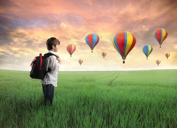 child carrying a backpack standing on a green meadow with hot-air balloons in the background