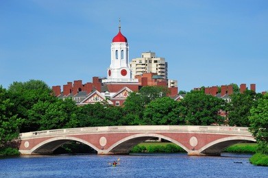 john w. weeks bridge and clock tower over charles river in harvard university campus in boston with trees, boat and blue sky.