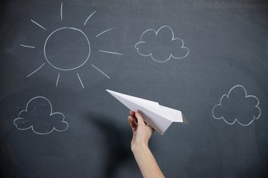 a female hand prepares to launch a paper airplane. blackboard with sky and sun on backgroud