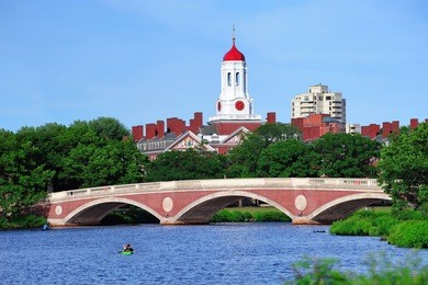 john w. weeks bridge and clock tower over charles river in harvard university campus in boston with trees, boat and blue sky.