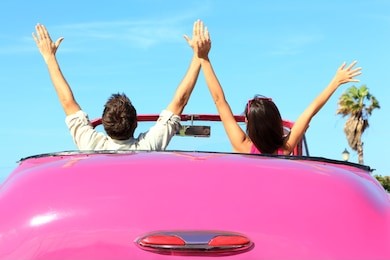 freedom - happy free couple in car driving in pink vintage retro car cheering joyful with arms raised. friends going on road trip travel on summer day under sun blue sky.