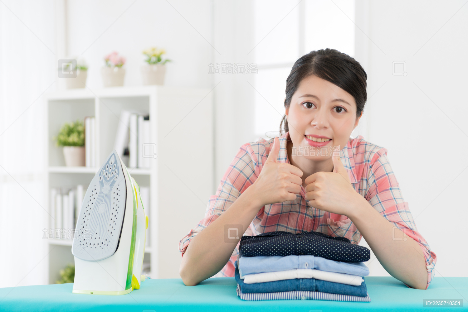 微笑幸福的女人管家熨烫在家的家庭服装和成品折叠面对镜头显示竖起大拇指的手势。