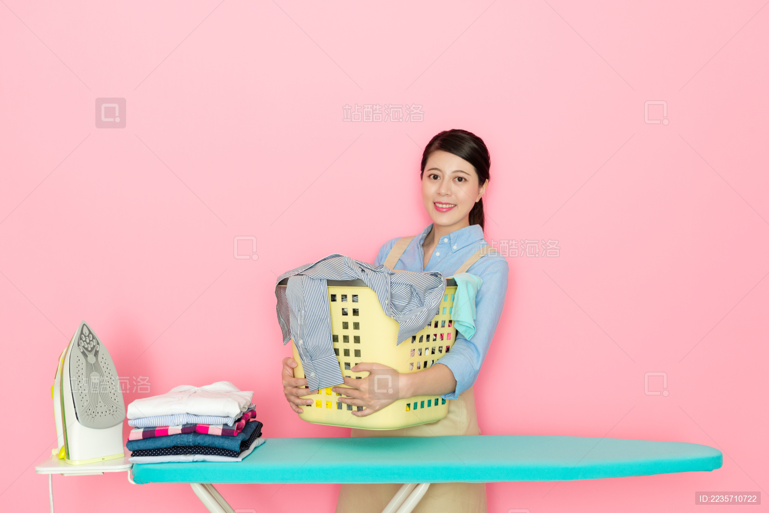 微笑优雅的家庭主妇拿着篮子 在粉红色的背景许多干净的衣服站在看着相机准备熨烫。