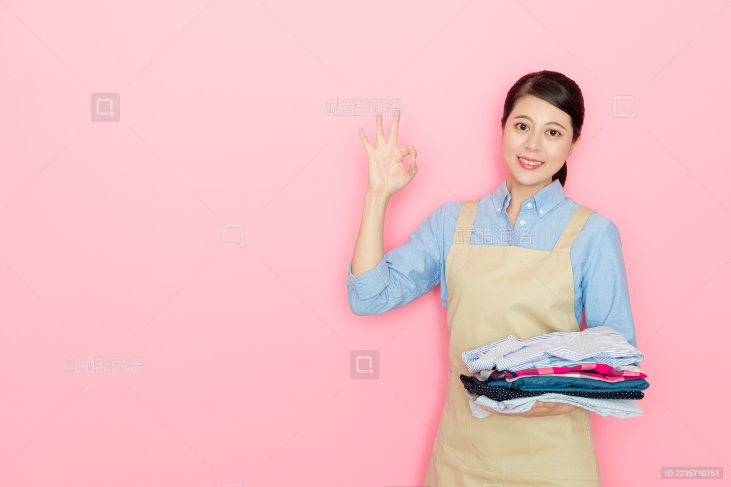 美丽快乐的女职工女仆拿着整齐的服装堆站在粉红色的背景 看着相机出OK手势。
