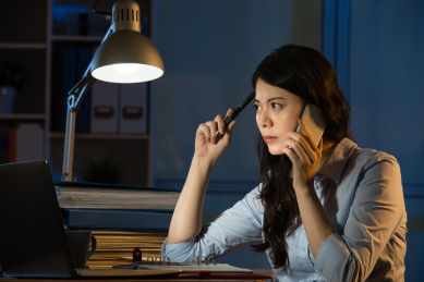 亚裔女商人在谈论智能手机加班深夜。在室内办公背景