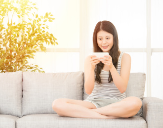 美丽的女人玩智能手机应用程序的游戏。多种族的女孩的短信应用，而坐在沙发上。亚洲高加索模型。