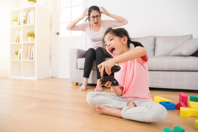 幸福的小女孩抱着孩子控制器玩视频游戏在客厅木地板有许多五颜六色的玩具，而她的母亲生气很生气显示在沙发上疯狂的情感。