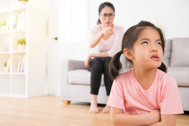 亚洲妈妈认真地坐在家里的沙发上愤怒指向的背面的小女孩不耐烦和不快乐的表达，而孩子会犯错。