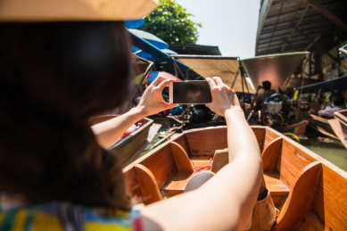 女性旅客使用移动手机采取浮动的市场来看风景的照片在泰国旅游坐在江上舟的后视图照片。
