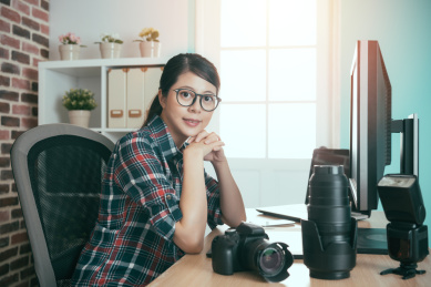 自信成功的商业摄影师妇女人权坐在编辑办公桌看着镜头微笑准备工作润饰图片。