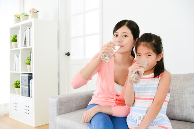 微笑的年轻母亲在客厅一起和饮用水看着镜头坐在沙发上沙发的小女儿。