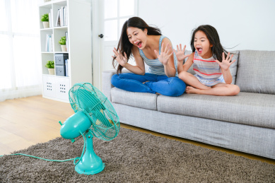 年轻的母亲与可爱的小女孩坐在沙发上沙发上，让笑冒充打电风扇享受着凉爽的风。