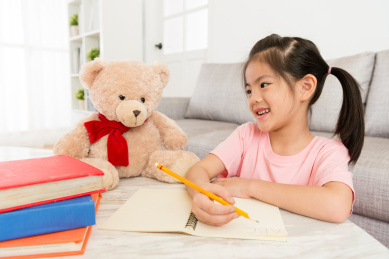 有吸引力的微笑的女学生的孩子与她最喜欢的玩具熊坐在客厅时，她写家庭作业在家里学习。