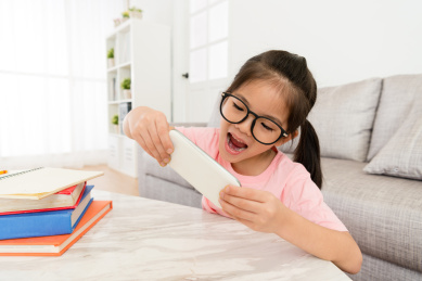 开朗可爱的小女孩使用移动智能手机玩在线视频游戏时，她长假期间在家里完成作业。