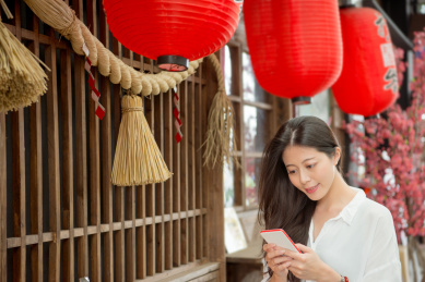 年轻女孩拿着移动手机搜索日本茶叶信息在红灯笼装饰的模糊日本文本意义的庆祝站在传统的日本茶的建筑。