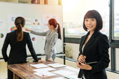 相信漂亮的女商人面对相机时，使用背景白板会议的她的同事微笑。