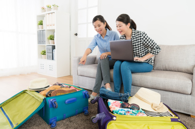 微笑的美闺蜜用笔记本电脑检查行李，手提箱指向确认在家里准备行李坐在沙发上沙发上。