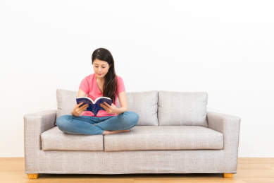 美丽的女学生坐在客厅沙发上，认真学习，认真度假阅读本书的白色背景上的木地板。
