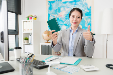 面带微笑的漂亮女人Manager显示与护照在看相机时，她坐在旅行社办公室竖起大拇指的手势。