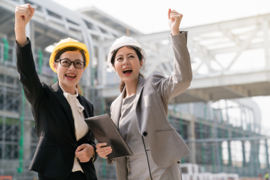 两个亚洲女工程师举手欢呼起来为他们在建筑物竞争中取胜。他们的笑容展现自己的幸福。在建筑工地背景。