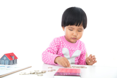 白背景前小女孩正在摆弄桌子上的欧元硬币以及桌子上还有计算器和小房子模型