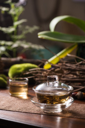 透明玻璃复古盖碗茶冲泡绿茶的场景，背景是原木桌面和绿色植物