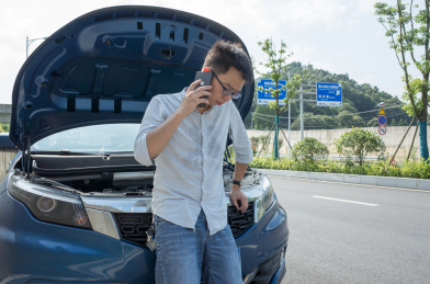 远处路边停着一辆坏掉的汽车，近处放着三角警告牌，一个男青年正在车前面打电话求助