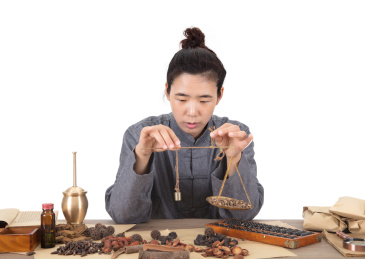 年轻的女性中医大夫在放满各种药材及用具的桌子前使用药秤称药