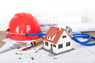 工程图纸和图纸上一个带着听诊器正给一个小房子模型听诊的安全帽和其它相关工具