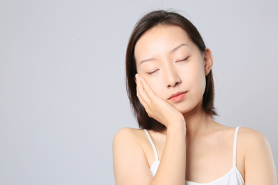 一位亚裔女孩站在纯色背景墙前托着脸颊闭起双眼