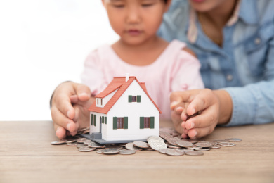 年轻妈妈和女儿用手呵护着美元硬币上放着的小房子模型