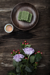 珊瑚红小杯里面盛满了祁眉红茶茶汤，搭配绿色龙井茶糕，朱红色紫砂小壶，下面几朵鲜花