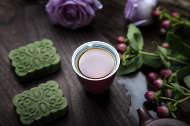 珊瑚红小杯里面盛满了祁眉红茶茶汤，搭配绿色龙井茶糕，朱红色紫砂小壶，周围几朵鲜花