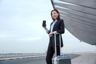 一位身穿正装面带笑容的女白领带着行李箱站在路旁展示手机屏幕