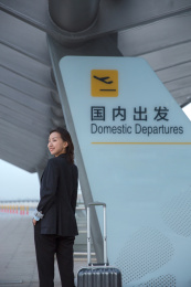 一位商务女士站在国内出发牌边回头微笑身边有一个拉杆箱
