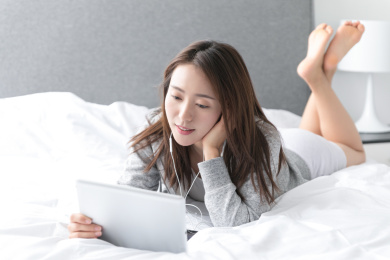 一名年轻女性趴在床上使用平板电脑听音乐