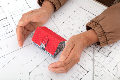 一双老年人的手呵护着平面图纸上的一个小房子模型