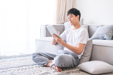 一名年轻的亚洲男子坐在客厅地毯上使用平板