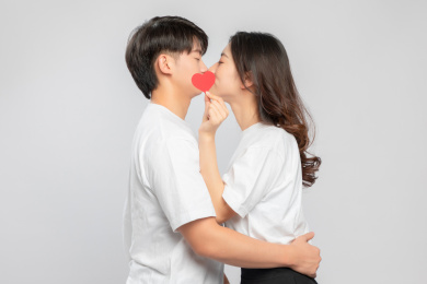 灰色背景里，手里拿着爱心纸片正在亲吻的亚洲年轻情侣