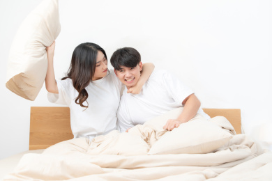 一对亚洲年轻情侣坐在床上正在拿着枕头玩耍，非常快乐的样子