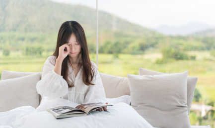 年轻长发女生坐在床上一边看书一边揉眼睛