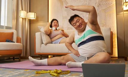 肥胖的青年人吃着汉堡看着伴侣健身
